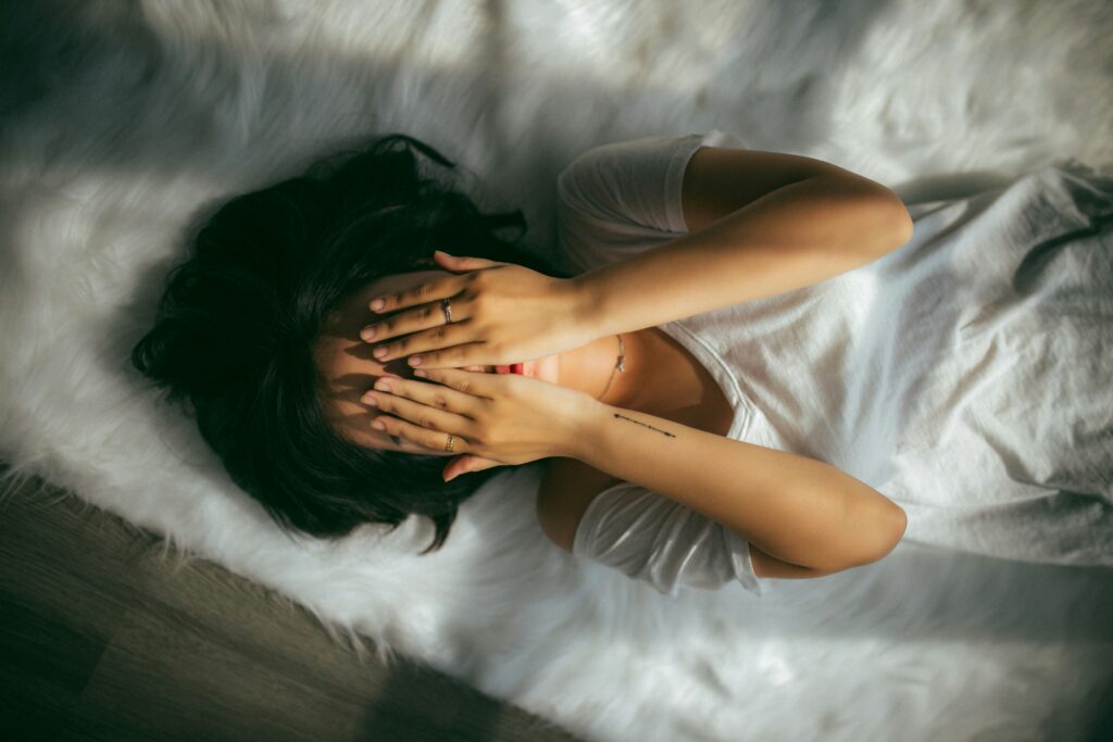 Vrouw met handen voor ogen op bed, symbool van moeheid, overbelasting en moeite met grenzen stellen.